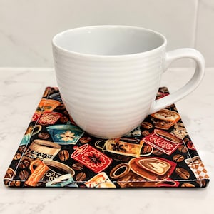 May include: A white ceramic mug with horizontal ridges sits on a square fabric coaster. The coaster features a black background with a repeating pattern of colourful coffee cups, mugs, and scattered brown coffee beans throughout the design.