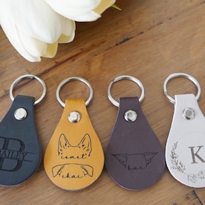 May include: Four leather keychains with different designs. The first keychain is black with a silver letter B and the name Bailey. The second keychain is tan with a dog silhouette and the name Comet. The third keychain is brown with a dog silhouette and the name Bal. The fourth keychain is white with a silver letter K and a floral design.
