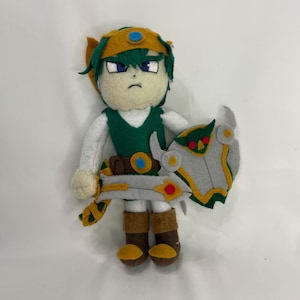 Puede incluir: Muñeca de fieltro hecha a mano de un personaje con pelo verde, túnica verde y botas marrones. La muñeca sostiene un escudo y una espada grises. La muñeca tiene una expresión seria y lleva una diadema dorada y azul.