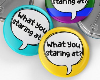 May include: Four round button pins with the text "What you staring at?" in a speech bubble. The buttons are in the colors blue, yellow, rainbow, and silver.
