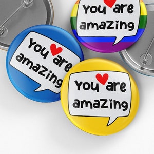 May include: A collection of round pinback buttons with the message "You are amazing" in a speech bubble. The buttons are in blue, yellow, and rainbow colors, with a red heart. The backs of the buttons are silver.
