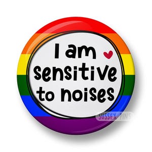 May include: A circular badge featuring a rainbow pride flag border. The badge displays the text "I am sensitive to noises" in black, with a small red heart. The words "Sussex Pins" are at the bottom.