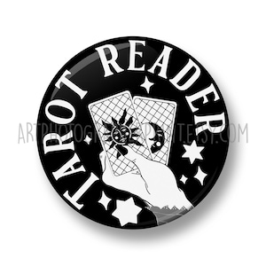 May include: Black and white button with the text "Tarot Reader" in a circle around a hand holding two tarot cards. The cards have a sun and moon design.