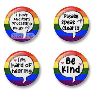 Può includere: Quattro bottoni rotondi con bordi arcobaleno e fumetti bianchi. I bottoni presentano le scritte: "I have Auditory Processing Issues", "Please speak clearly", "I'm hard of hearing" e "Be Kind". Il testo è nero.