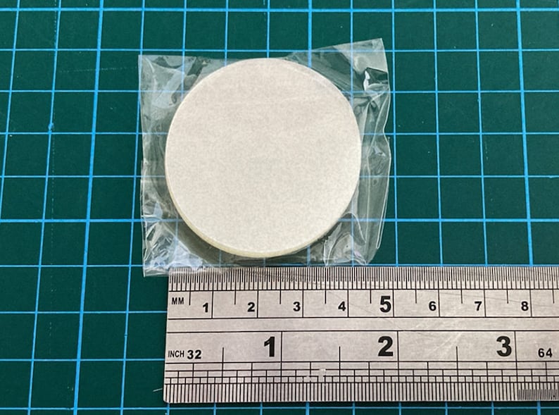 K&ouml;nnte beinhalten: Eine Packung runde, cremefarbene Papierfilter, etwa 3,8 cm im Durchmesser, ist in einem durchsichtigen Plastikbeutel versiegelt. Die Filter liegen auf einer gr&uuml;nen Rasteroberfl&auml;che mit einem Lineal zur Ma&szlig;stabsbestimmung.