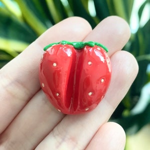 以下が含まれることがあります： 白いドットが付いた赤と緑の粘土製のイチゴチャーム。チャームはハート型で、緑色の茎と葉が付いています。