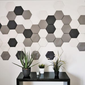 May include: A white wall with a geometric pattern of hexagonal tiles in shades of gray and black. The tiles are arranged in a staggered pattern, creating a modern and stylish look. Two potted plants and a candle are on a black table in front of the wall.