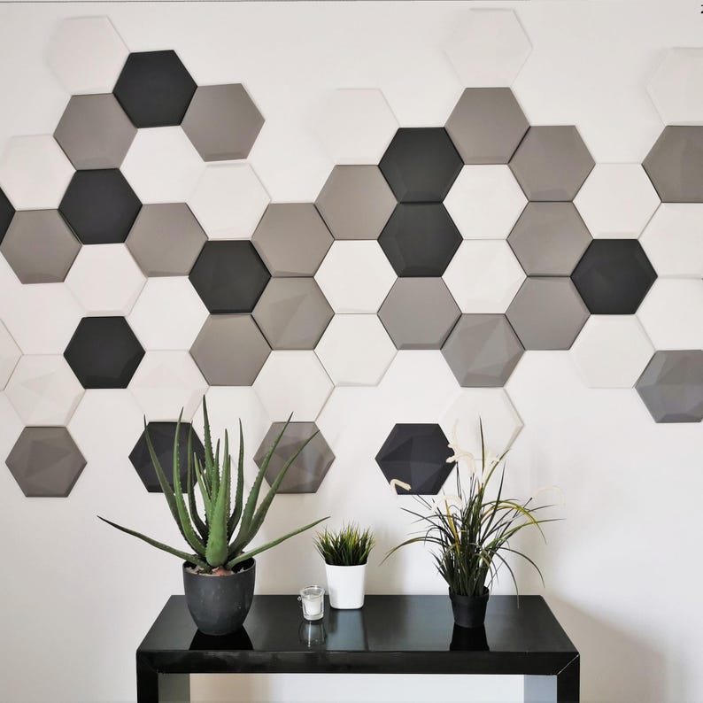 May include: A white wall with a geometric pattern of hexagonal tiles in shades of gray and black. The tiles are arranged in a staggered pattern, creating a modern and stylish look. Two potted plants and a candle are on a black table in front of the wall.