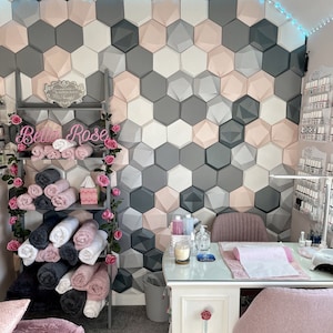 Peut inclure: Une table blanche avec un mur hexagonal rose et gris derrière elle. La table a un dessus blanc et des pieds blancs. Il y a une chaise rose au premier plan. La table a un bol blanc avec une bougie à l'intérieur, une bouteille de désinfectant pour les mains et une bouteille de lotion. Il y a une serviette blanche sur la table. Le mur a une pancarte qui dit "Bella Rose".