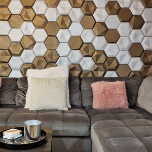 May include: A gray sectional sofa with two pillows, one white with a patterned design and one pink with a fluffy texture. The sofa is in front of a wall with a geometric pattern of white and gold hexagonal tiles.