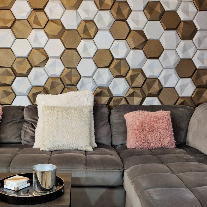 May include: A gray sectional sofa with two pillows, one white with a patterned design and one pink with a fluffy texture. The sofa is in front of a wall with a geometric pattern of white and gold hexagonal tiles.
