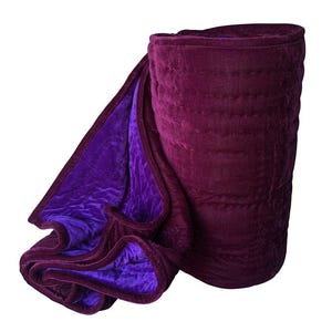 Peut inclure: Un plaid en velours moelleux présentant un extérieur bordeaux profond et un intérieur violet vif. Le tissu présente des surpiqûres horizontales sur toute la surface, créant une texture avec un drapé souple et plié sur un côté.
