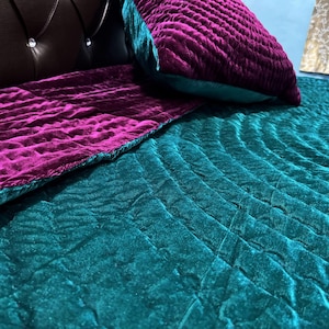 Op de afbeelding: Close-up van een sprei en kussens in teal en paars fluweel. De sprei heeft een gewatteerd patroon en de kussens zijn rechthoekig. De fluwelen stof heeft een zachte, luxueuze uitstraling.