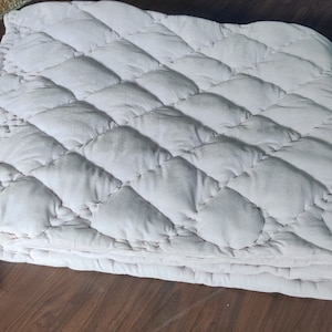 Könnte beinhalten: Eine weiche, hellrosa gesteppte Decke mit Rautenmuster und weißen Quasten.