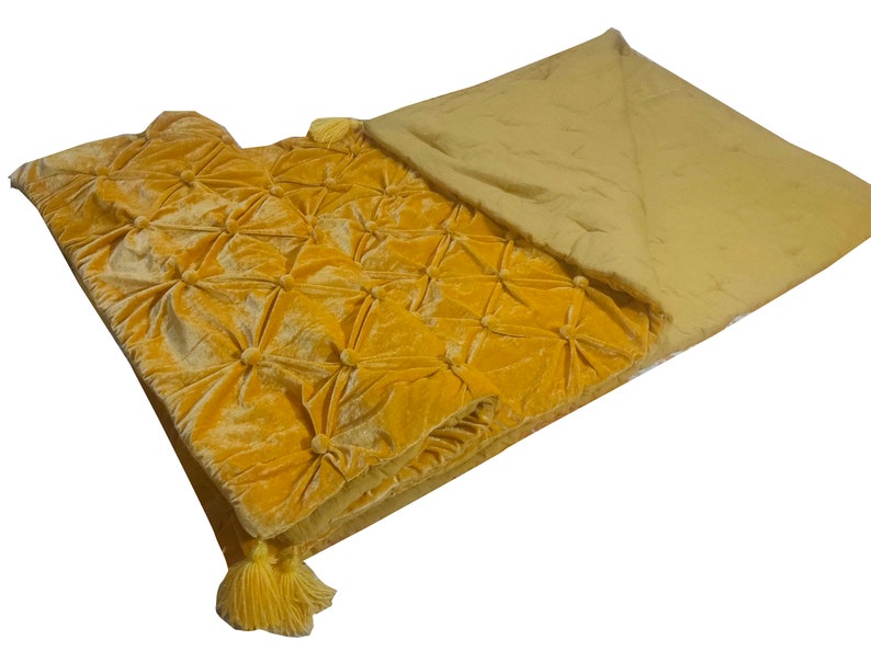 K&ouml;nnte beinhalten: Eine gelbe Samtdecke mit einem gesteppten Muster und Quasten. Die Decke ist in der H&auml;lfte gefaltet und die Unterseite ist einfarbig gelb.