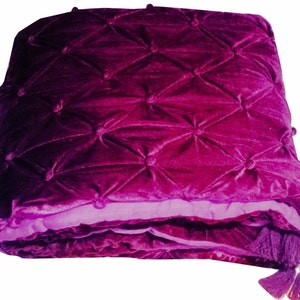 K&ouml;nnte beinhalten: Eine lilafarbene Samtdecke mit Steppmuster und Quasten. Die Decke hat einen Farbverlauf von einem dunkleren Lila zu einem helleren Lila.