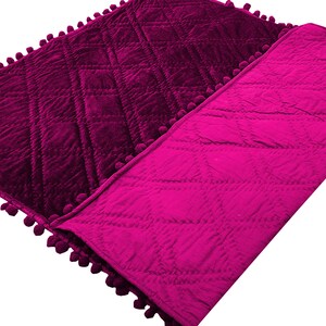 Może przedstawiać: R&oacute;żowo-magenta pikowana kołdra z ozdobnym obszyciem z pompon&oacute;w. Kołdra jest złożona na p&oacute;ł, odsłaniając pikowany wz&oacute;r po obu stronach.