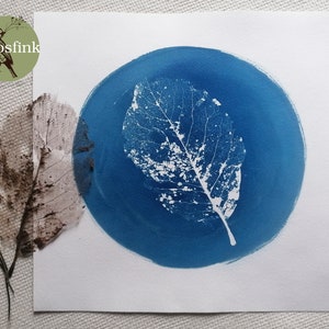 May include: A white leaf print on a blue circular background. The print is a cyanotype, a photographic printing process that uses light-sensitive iron salts.