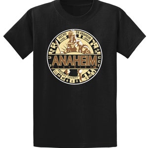 May include: Black t-shirt with a brown and gold circular graphic featuring a warrior holding a spear and shield. The graphic says "Hecho en Anaheim" and "Anaheim Clothing Orange County".
