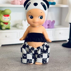 May include: A small figurine wearing a white panda-themed headpiece with black ears and eyes. The figure is dressed in a black strapless top and loose-fitting pants featuring a black and white checkered pattern with a textured, speckled finish.