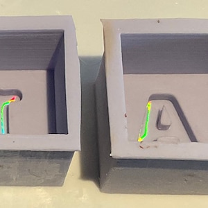 Può includere: Due stampi in silicone grigi per realizzare oggetti in resina. Gli stampi sono di forma quadrata e hanno le lettere "T" e "A" all'interno.