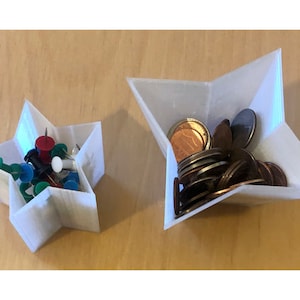 Puede incluir: Dos contenedores blancos en forma de estrella, uno más grande que el otro. El contenedor más grande está lleno de monedas, mientras que el contenedor más pequeño contiene chinchetas de varios colores.