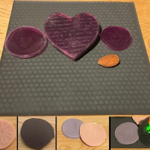 Op de afbeelding: Een set van acht paarse en roze rubberen stempels met verschillende ontwerpen, waaronder een hart, een hand met een vredesteken en een bloem. De stempels liggen op een grijs, getextureerd oppervlak.