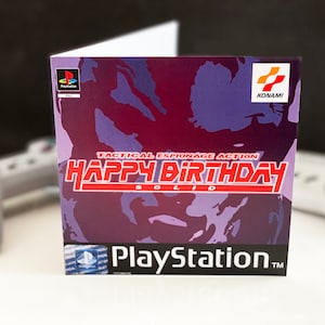 May include: A birthday card featuring a purple and red camouflage pattern with the text "Tactical Espionage Action Happy Birthday Solid" and the Konami logo. The card also has the PlayStation logo.
