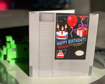 Op de afbeelding: Een grijze en zwarte Nintendo Entertainment System-spelcartridge met een gepixelde verjaardagstaart en een rode ballon aan de voorkant. De tekst "Happy Birthday!" staat op de voorkant van de cartridge.