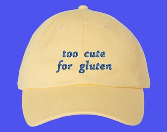 May include: A pale yellow baseball cap featuring the embroidered text "too cute for gluten" in a dark blue font. The cap has a structured crown, a curved brim, and a button at the top center. The design is centered on the front panels of the hat.