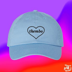 Op de afbeelding: Een lichtblauwe baseballpet met een geborduurde zwarte hartomtrek op het voorpaneel. Binnenin het hart is het woord thembo geborduurd in een zwart, kleine letters, schreefloos lettertype. De pet heeft een gebogen klep en een gestructureerde kroon.