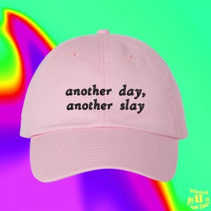 Op de afbeelding: Een lichtroze honkbalpet met de geborduurde zwarte tekst "another day, another slay" gecentreerd op het voorpaneel. De pet heeft een gestructureerde kroon, een gebogen klep en een knoop aan de bovenkant, wat zorgt voor een strak ontwerp.