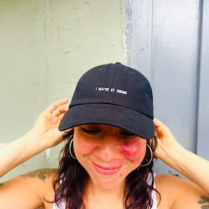 May include: A black baseball cap featuring the white embroidered text I HATE IT HERE centered on the front panel. The cap has a structured crown, a curved brim, and a button at the top, constructed from a solid black fabric material.