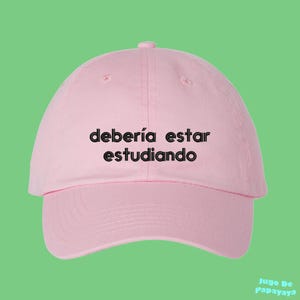 以下が含まれることがあります： フロントパネルの中央に黒い刺繍で「debería estar estudiando」というテキストが施されたライトピンクの野球帽。構造化されたクラウン、カーブしたつば、上部のボタンが特徴で、シンプルで洗練されたデザインに仕上がっています。