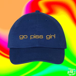 Puede incluir: Una gorra de béisbol azul marino con el texto bordado "go piss girl" en color dorado en el panel frontal. La gorra tiene una corona estructurada, una visera curva y un botón en la parte superior, mostrando un diseño sencillo.