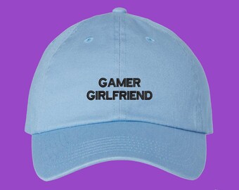 May include: A light blue baseball cap featuring the text GAMER GIRLFRIEND embroidered in black thread across the front center. The cap has a structured crown, a curved brim, and a button at the top, presenting a simple and clean design.