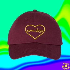 May include: A maroon baseball cap with a yellow heart embroidered on the front. The heart has the words "corn dogs" inside.
