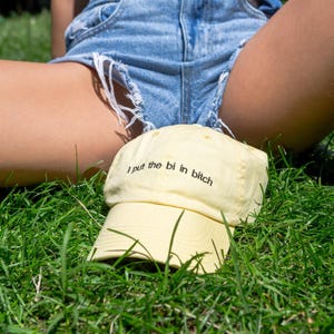 Peut inclure: Une casquette de baseball jaune pâle avec le texte brodé en noir "I put the bi in bitch" sur le panneau avant. La casquette présente une visière incurvée et une calotte structurée, fabriquée dans un tissu uni avec des coutures assorties.