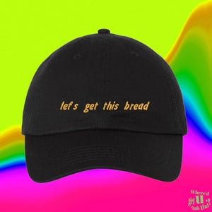 May include: A black baseball cap featuring the embroidered text "let's get this bread" in a yellow cursive font across the front panel. The cap has a structured crown, a curved brim, and a button at the top center of the dome.