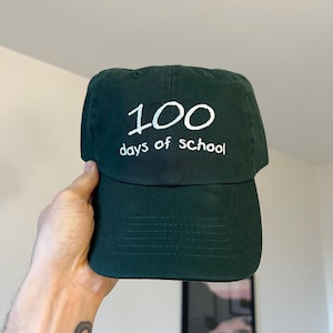 May include: A dark green baseball cap featuring white embroidered text that reads 100 days of school. The cap has a structured crown, a curved brim, and a classic six-panel design with ventilation eyelets on the top of the fabric crown.