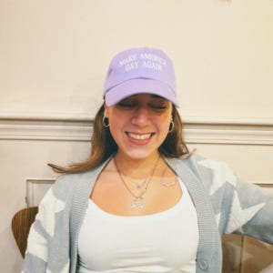 May include: A person wearing a light purple baseball cap with the text "Make America Gay Again" embroidered in white. The person is smiling and wearing a light gray cardigan with a white pattern.
