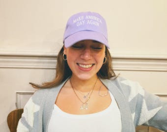 Könnte beinhalten: Eine Person trägt eine helllila Baseballkappe mit dem Schriftzug "Make America Gay Again", der in Weiß gestickt ist. Die Person lächelt und trägt einen hellgrauen Cardigan mit weißem Muster.