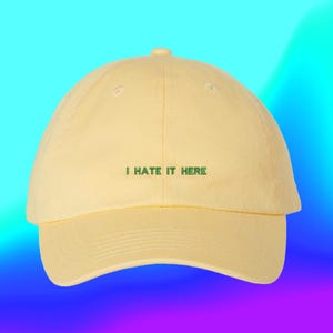 May include: A pale yellow baseball cap featuring the embroidered text I HATE IT HERE in dark green capital letters centered on the front panel. The cap has a structured crown, a curved brim, and a button at the top of the dome.