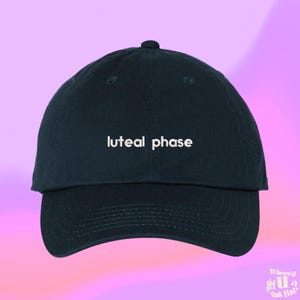 以下が含まれることがあります： 紺色のベースボールキャップ。白い文字で「luteal phase」と刺繍されています。キャップはカーブしたつばと丸いトップが特徴です。背景はピンクと紫のグラデーションです。