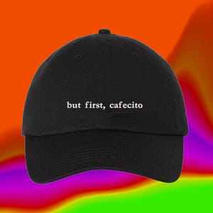 May include: A black baseball cap with a curved brim and a structured crown. The front of the cap features white embroidered text that reads, but first, cafecito. The design is centered on the front panel, showcasing a clean and simple aesthetic.