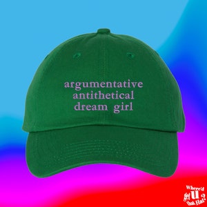 Peut inclure: Une casquette de baseball verte avec le texte "argumentative antithetical dream girl" brodé en rose.