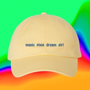 Può includere: Un cappello da baseball giallo con il testo "manic pixie dream girl" ricamato in blu sul davanti.