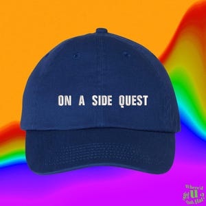 May include: A navy blue baseball cap featuring the white embroidered text ON A SIDE QUEST centered on the front panel. The cap has a structured crown, a curved brim, and a button at the top. The design is simple, clean, and functional.