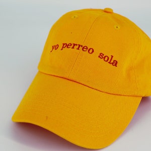 May include: A yellow baseball cap with the phrase "yo perreo sola" embroidered in red.