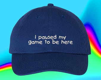 May include: A navy blue baseball cap featuring the white embroidered text "i paused my game to be here" centered on the front panel. The cap has a structured crown, a curved brim, and a button at the top, presenting a classic six-panel design.
